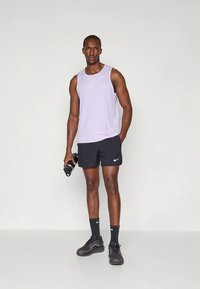 Vaaleanpurppura urheilullinen hihaton paita pyöreällä kaula-aukolla ja musta logo, yhdistetty mustiin shortseihin ja tummiin lenkkareihin, kädessä vesipullo.