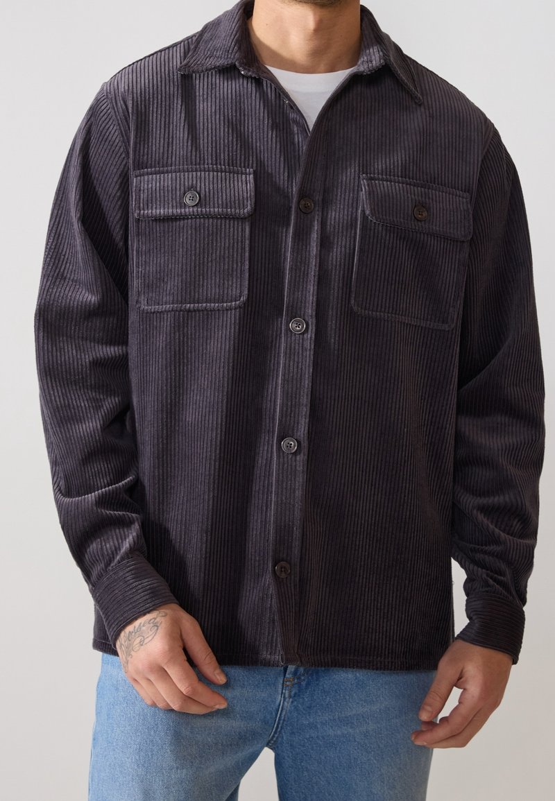 Camicia in corduroy grigio scuro con due tasche sul petto, chiusura a bottone e colletto a punta. Presenta una texture rigata e maniche lunghe.
