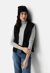 Frau trägt eine schwarze Mütze, eine schwarze ärmellose Strickjacke über einem grauen Rollkragenpullover und blaue Jeans, steht vor einer weißen Wand.