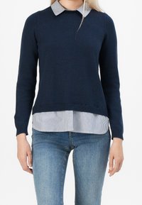 Pull en maille bleu marine avec un col rond, doté d'un col rayé. Porté sur une chemise blanche à fines rayures bleues. Associé à un jean bleu.