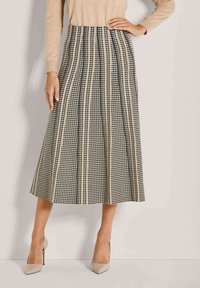 Beige und schwarze kariert midi Rock, getragen mit beigem Langarmoberteil und beigen High-Heels, Modell steht mit einer Hand auf der Hüfte.