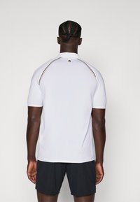 T-shirt de sport à manches courtes, blanc, avec un col haut, présentant des rayures contrastantes noires et dorées le long des épaules. Texture lisse.