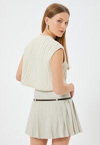 Jersey de crema de punto sin mangas y recortado con un patrón texturizado, combinado con una falda plisada ligera y un cinturón negro.