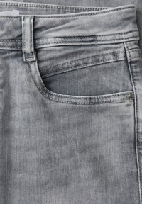 Close-up van een vervaagde grijze spijkerbroekzak met zichtbare riemlus, stiknaden en metalen klinknagel aan de rechterkant.