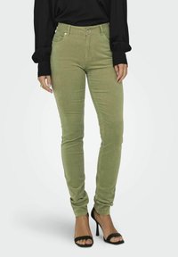 Jeans skinny verts en tissu texturé avec une taille mi-haute, fermeture à bouton et poches avant. Associés à un haut noir à manches longues et des talons.