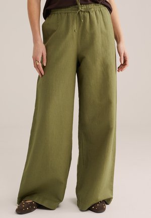 Žena stojaca pred neutrálnym pozadím, oblečená v olivovo zelenej širokých nohaviciach s nastaviteľným pásom a tmavohnedých plochých topánkach so cvočkami.