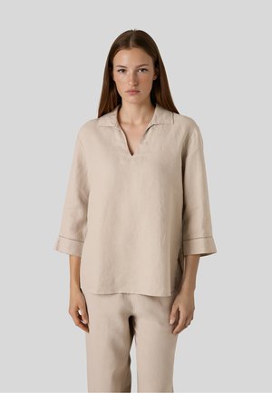 Kvinde med glat brunt hår iført løs beige top med 3/4 ærmer og matchende bukser, står mod en ensfarvet grå baggrund.