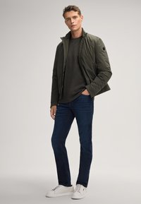 Dunkelgrüne gepolsterte Jacke mit hohem Kragen, getragen über einem schwarzen Hemd, kombiniert mit blauen Jeans und weißen Sneakers. Glatte Textur, minimalistisches Design.