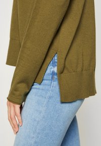 Personne portant un jean bleu clair et un pull vert olive avec des fentes latérales, vue de la mi-cuisse à la taille sur le côté gauche.