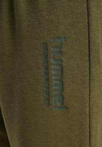 Olivenfarvede sportswear-bukser med ribbet tekstur, der har et mørkegrønt "hummel"-logo og teksten "SPORTSWEAR" langs sidesømmen.