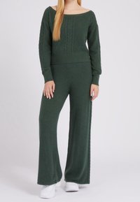 Femme portant un pull en maille torsadée vert foncé et un pantalon large assorti, avec des baskets blanches, debout devant un fond uni.