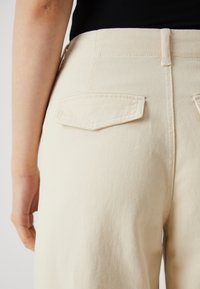 Pantalon en coton beige avec taille haute, deux poches arrière à rabat et tissu texturé. Couture visible le long des coutures.