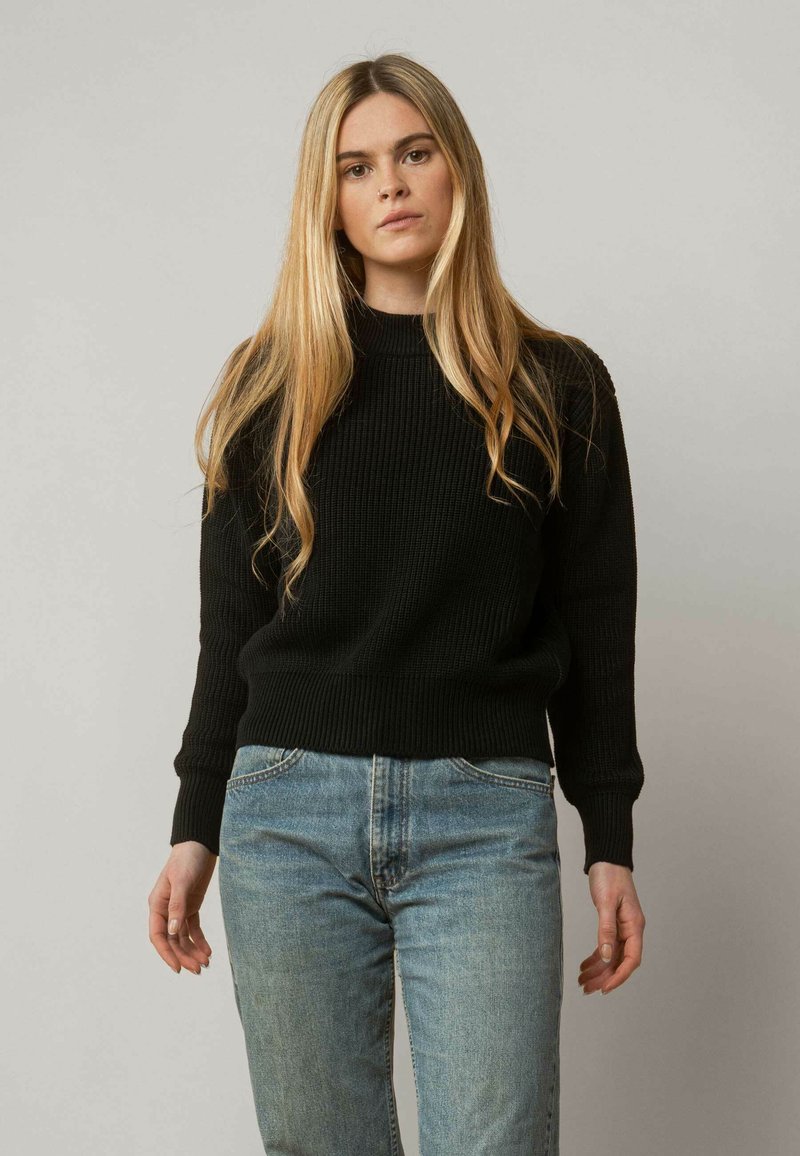 Schwarzer gerippter Strickpullover mit Rundhalsausschnitt und langen Ärmeln, kombiniert mit hellblauen Denim-Jeans. Schlichtes und figurnahes Design.