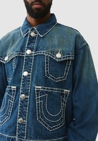 Veste en denim bleu avec col à revers, boutons argentés et deux poches avant avec couture contrastée et détails de design distinctifs.