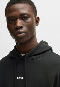 Czarna bluza z kapturem wykonana z miękkiego materiału, z kapturem na sznurku i białym logo "BOSS" umieszczonym na środku klatki piersiowej. Zbliżenie na portret.