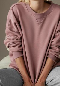 Persoon die een losse, mauve sweatshirt met opgerolde mouwen en grijze broek draagt, zittend met de handen op de dijen rustend.