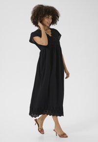 Black cotton dress with short sleeves, scalloped hem, and eyelet lace detailing. Model wearing brown heeled sandals.