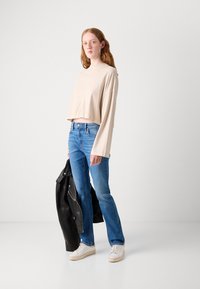 Beige cropped langermet topp, blå rettsnitt jeans, hvite sneakers og svart skinnjakke holdt i hånden. Glatt stoff og enkelt design.