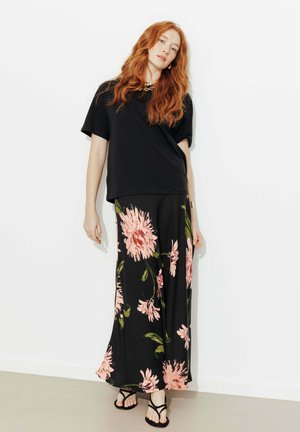 Femme aux cheveux roux portant un t-shirt noir, une jupe longue noire à motifs floraux, et des sandales noires, debout contre un mur blanc.