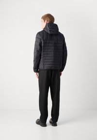 Casaco preto acolchoado com capuz, apresentando uma textura acolchoada e costuras horizontais. Combinado com calças pretas e sapatos desportivos pretos.