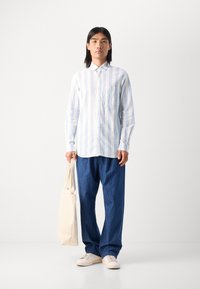 Chemise boutonnée à rayures bleu clair et blanches avec manches longues, associée à un pantalon en denim bleu ample et un sac fourre-tout en toile crème.