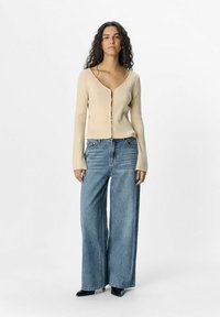 Cardigan beige a coste con scollatura a V e chiusura a bottoni, abbinato a jeans in denim blu a gamba larga con vita alta. Scarpe nere con tacco.