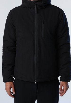 Homme portant une veste matelassée noire avec capuche, fermeture éclair frontale et poche poitrine zippée, debout devant un fond clair.