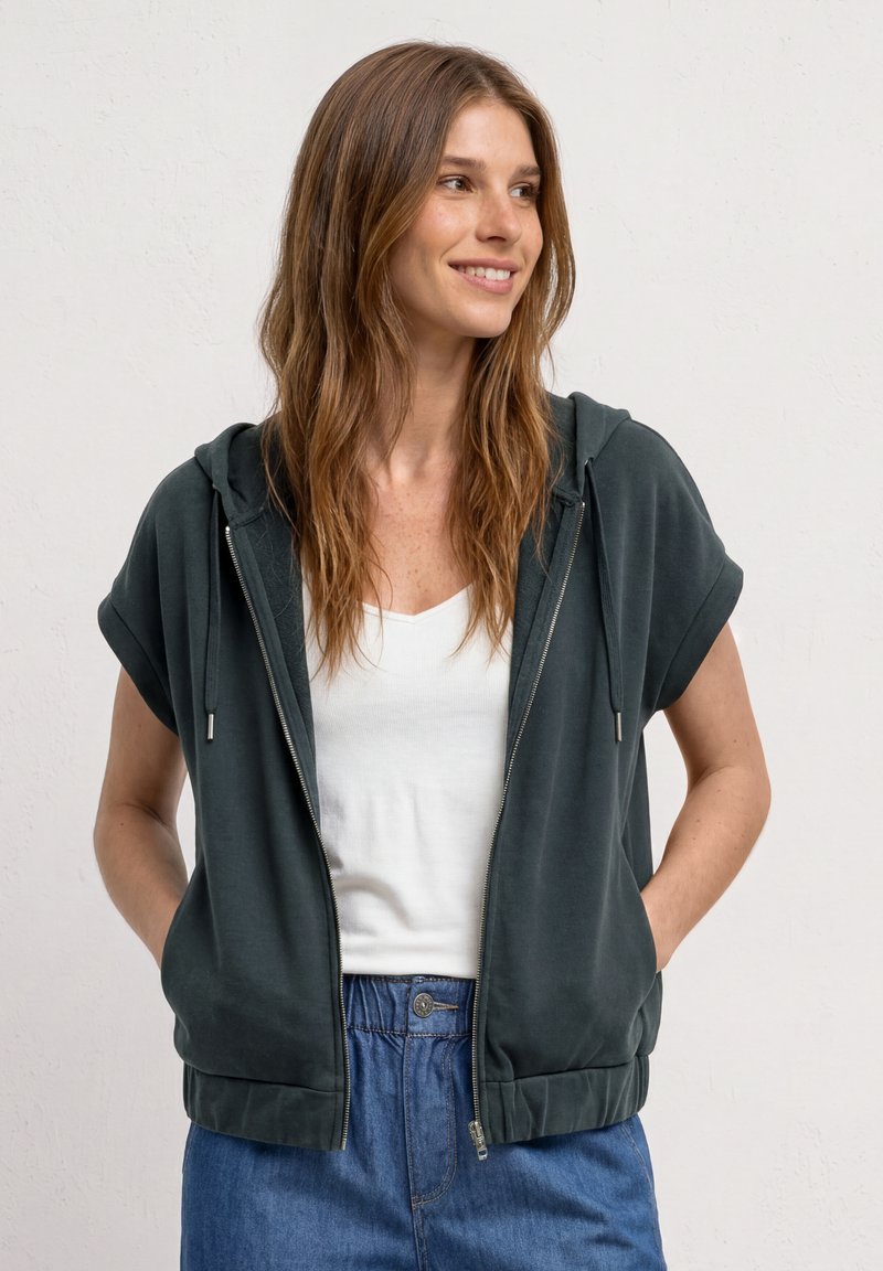 Femme souriante aux longs cheveux bruns portant un sweat à capuche vert foncé à manches courtes zippé, un débardeur blanc et un jean bleu, les mains dans les poches.