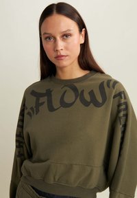 Sudadera verde oliva con un diseño cropped y cuello redondeado. Detalles en texto negro "FLOW" y "FREDDY" en las mangas. Textura suave.