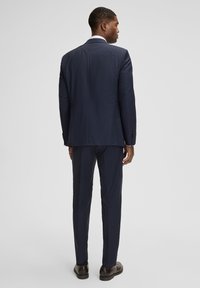 Marineblauer, maßgeschneiderter Anzug mit dezentem Karomuster, bestehend aus einem Sakko mit Einzelreißverschluss, Slim-Fit-Hosen und dunklen Anzugschuhen.