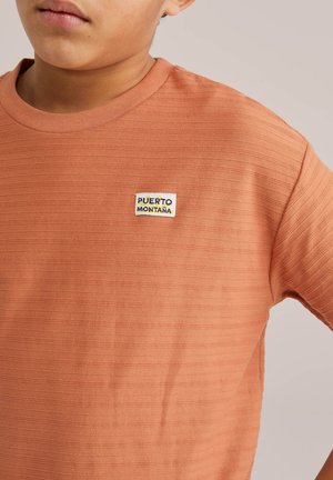 Kind trägt strukturiertes oranges Kurzarmshirt mit kleinem "Puerto Montaña"-Label auf der Brust, beiger Hintergrund.