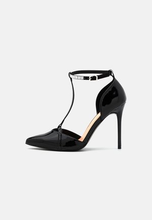 Czarny lakierowany but na wysokim obcasie z ostrym noskiem, pasek wokół kostki, design w stylu T-strap oraz cienki obcas szpilka na jednolitym tle.