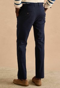 Donkerblauwe katoenen broek met een rechte pijp, voorzien van achterzakken en knoopdetails, gecombineerd met bruine instapschoenen.