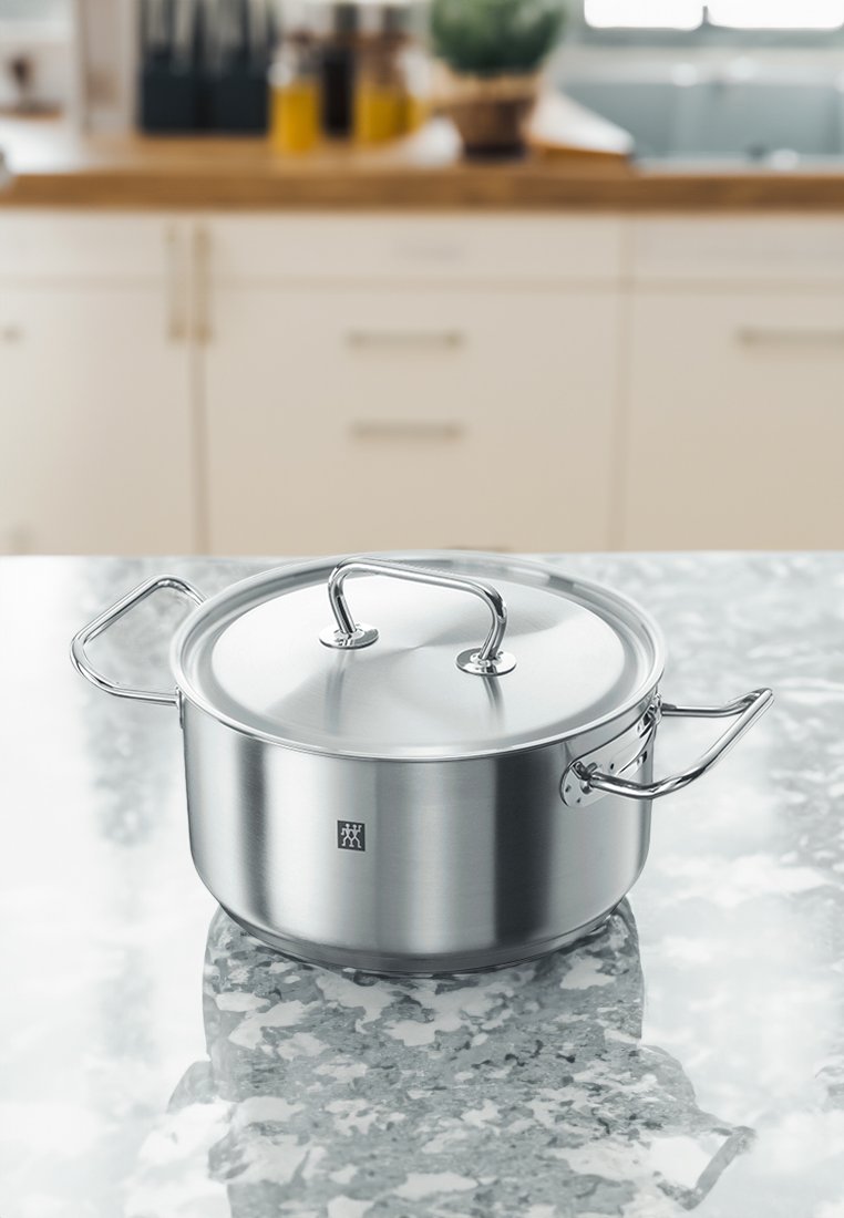 Casserole en acier inoxydable avec un couvercle en verre, de forme ronde, deux poignées en acier inoxydable, et un logo à l'avant, placée sur une surface en marbre.