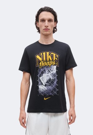 Homme portant un t-shirt noir Nike Hoops avec un motif de basketball et clôture en grillage, associé à un short de sport blanc.