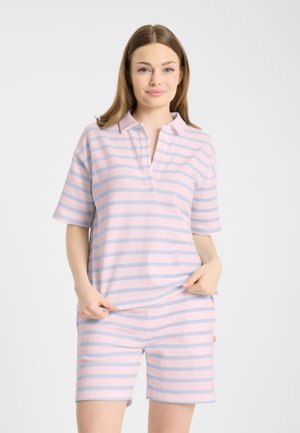 Młoda kobieta ubrana w różowo-niebieską w paski koszulkę z krótkim rękawem i pasujące krótkie spodenki, stojąca na jednolitym tle.