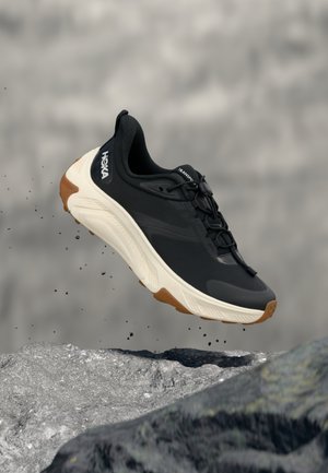 Schwarz-weißer Hoka-Laufschuh in der Luft über felsigem Gelände, mit fallenden Schmutzpartikeln um die Sohle, unscharfer grauer Hintergrund.