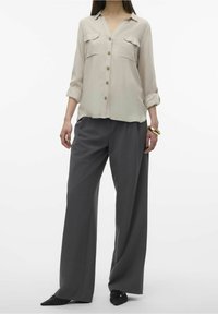 Blusa beige chiaro con un frontale con bottoni, due tasche sul petto e maniche arrotolate. Abbinata a pantaloni larghi grigio scuro e scarpe nere.