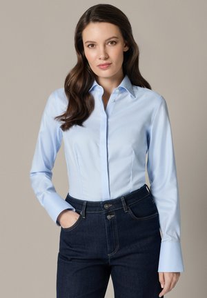 Kobieta z długimi brązowymi włosami, ubrana w jasnoniebieską koszulę zapinaną na guziki i ciemnoniebieskie jeansy, stojąca z jedną ręką w kieszeni.