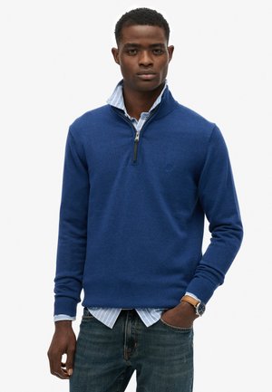 Mann trägt einen blauen Pullover mit Viertelreißverschluss über einem blau gestreiften Hemd mit Kragen, dazu Jeans und eine Armbanduhr, steht vor einem einfarbigen Hintergrund.