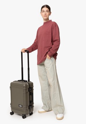 Maleta rígida de color verde oliva con asa extensible negra y ruedas. Colocada al lado de una persona que lleva puesta una camisa de manga larga color burdeos y jeans claros.