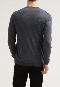 Mann in einem dunkelgrauen Langarmpullover und schwarzen Hosen, der mit dem Rücken zu einem schlichten, hellen Hintergrund steht.