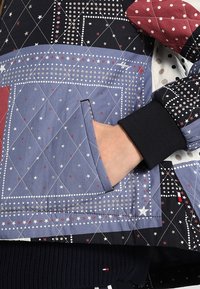 Veste matelassée avec des motifs géométriques en bleu marine, rouge et blanc. Comprend une poche latérale et des poignets côtelés. Tissu texturé avec des motifs d'étoiles.