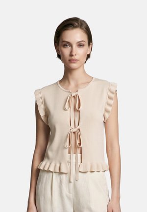 Giovane donna che indossa un top beige senza maniche con chiusure a fiocco sul davanti e bordi arricciati, abbinato a pantaloni chiari.