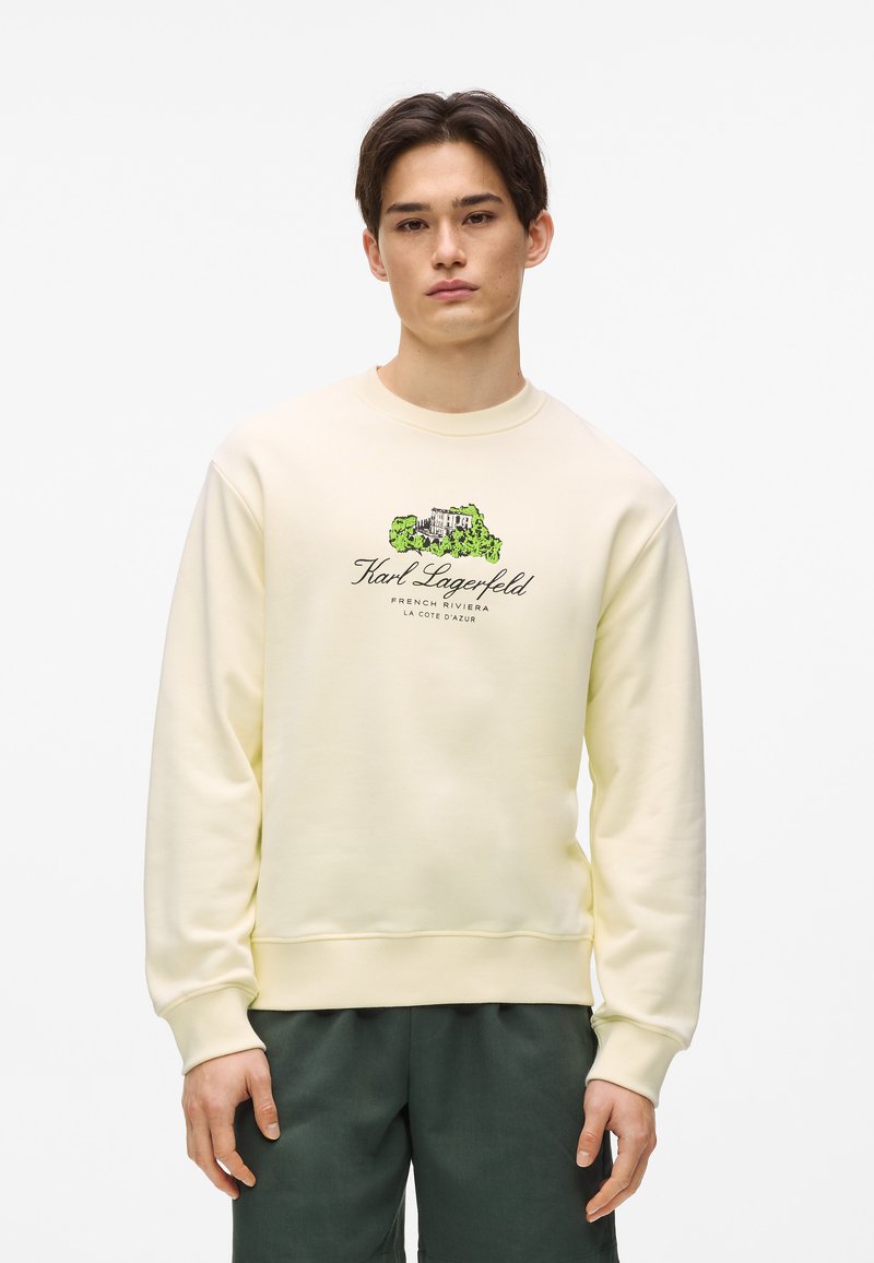 Sweat-shirt jaune clair en tissu doux avec col rond, poignets et taille côtelés. Présente un texte graphique et des illustrations vertes sur le devant.
