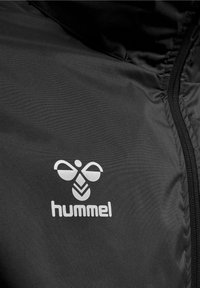 Tissu de veste noire avec un logo d’abeille stylisé blanc et le mot hummel imprimé en dessous près d'une fermeture éclair visible.