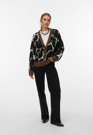 Kvinde iført en brun og sort cardigan med dyreprint, hvid skjorte, sorte bukser med vide ben, brune sko og en perlehalssmykke, stående mod en hvid baggrund.
