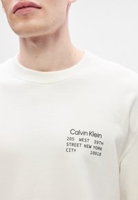 Λευκή μπλούζα με πλεκτό λαιμό ; διαθέτει μαύρο εκτυπωμένο κείμενο: "Calvin Klein 205 WEST 39TH STREET NEW YORK CITY 10018."