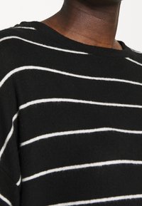 Schwarzer Strickpullover mit weißen horizontalen Streifen, rundem Halsausschnitt, weicher Textur und lässiger Passform. Nahaufnahme des Stoffdetails.