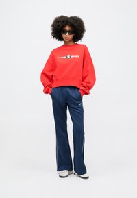 Červená nadrozmerná mikina s logom "NIKE SHOX", skombinovaná s námorníckymi športovými nohavicami s bielymi bočnými prúžkami a sivými teniskami. Čierne slnečné okuliare.